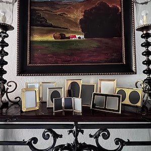 May include: A collection of various picture frames with gold and black finishes, arranged on a dark wooden surface. The frames are in different shapes and sizes, with some featuring a double frame design. A landscape painting hangs in the background.