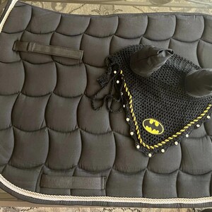 Puede incluir: Silla de montar acolchada negra para caballos con un logotipo de Batman en una capota a juego. La almohadilla tiene un ribete de cuerda blanca y plateada. La capota es negra con un ribete amarillo y blanco.