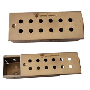 May include: Two rectangular stainless steel sterilization cases with perforated lids. Each lid features two rows of circular holes and is engraved with the text Germany Stainless CE. One case is closed, while the other is partially open.