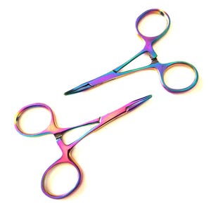 Puede incluir: Dos pinzas iridiscentes de color arcoíris con lazos para los dedos. Las pinzas tienen puntas afiladas y un mecanismo de resorte. La herramienta de metal refleja un espectro de colores, incluyendo púrpura, azul, verde y amarillo, sobre un fondo blanco.