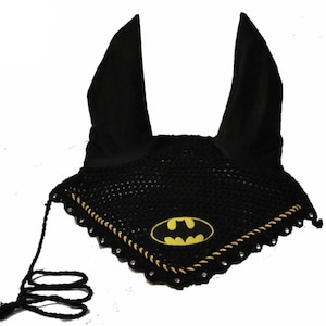 Op de afbeelding: Zwarte vliegenmuts voor paarden met een geel Batman-logo. De muts heeft oorbedekkingen en een gehaakte voorkant met een gouden rand en een zwart koord. Het Batman-logo staat in het midden aan de voorkant.