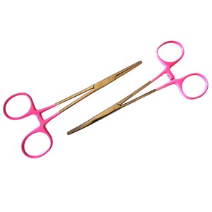 Puede incluir: Dos pinzas hemostáticas médicas. Los mangos son de color rosa brillante y las mandíbulas doradas. Una es más grande que la otra. Las pinzas están diseñadas para sujetar los vasos sanguíneos y detener el sangrado.
