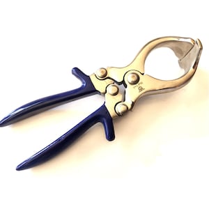 May include: A silver-colored metal tool with dark blue handles. The tool has a circular opening and a hinged design, suggesting it is used for gripping or clamping. The handles are curved for a comfortable grip.