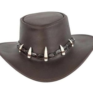 Könnte beinhalten: Ein brauner Lederhut mit breiter Krempe und rundem Oberteil. Der Hut hat ein dekoratives Band um den Ansatz des Oberteils, verziert mit weißen und braunen Akzenten. Ein stilvolles Accessoire.