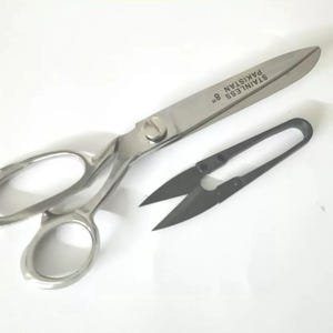 May include: A pair of silver scissors with a black handle and the text "STAINLESS 8" "PAKISTAN" on the blade. The scissors are on a white surface.