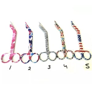 Puede incluir: Cinco pares de tijeras para cutículas con patrones coloridos. Las tijeras están dispuestas en una fila de izquierda a derecha, numeradas del 1 al 5. Cada par de tijeras tiene un patrón diferente, incluyendo rosa y blanco, azul y blanco, negro y blanco, rojo y blanco, y rojo, blanco y azul.