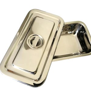 May include: Stainless steel rectangular tray with a matching lid. The lid features a central handle. The tray is designed with rounded corners and a polished, reflective surface.