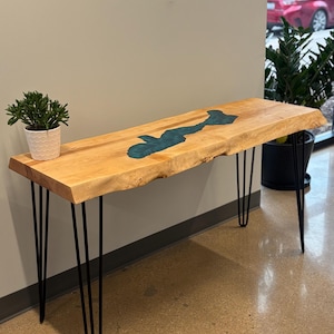 Puede incluir: Una mesa consola de madera con una tapa de borde vivo y un río de epoxi azul verdoso. La mesa está sostenida por cuatro patas de horquilla negras.