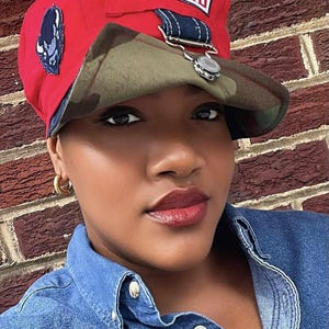 May include: Red cap with a camouflage brim and the word "HOWARD" in a white rectangle. The cap has a small patch on the side and a silver-colored decorative element. The person is wearing a denim shirt.