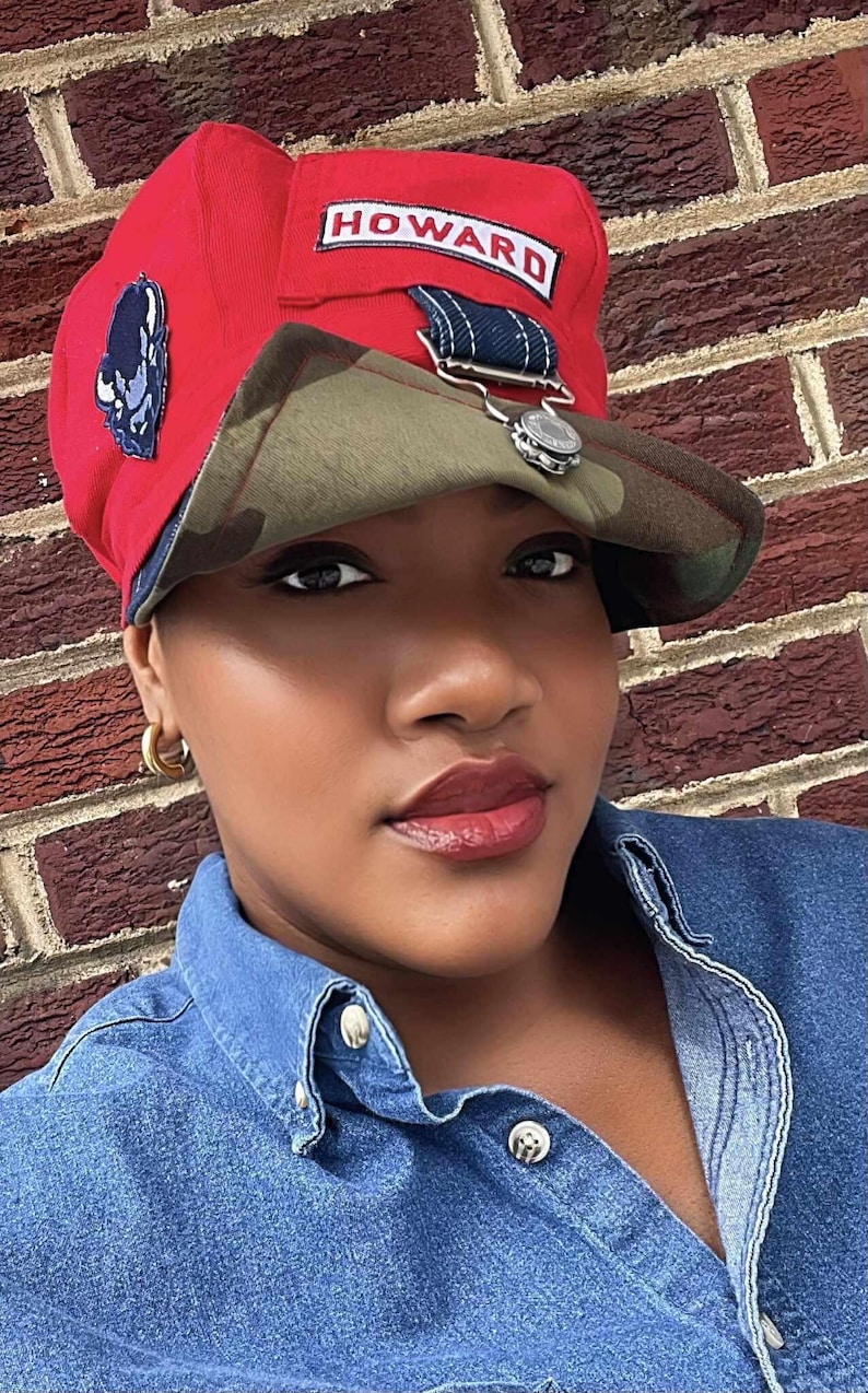 May include: Red cap with a camouflage brim and the word "HOWARD" in a white rectangle. The cap has a small patch on the side and a silver-colored decorative element. The person is wearing a denim shirt.
