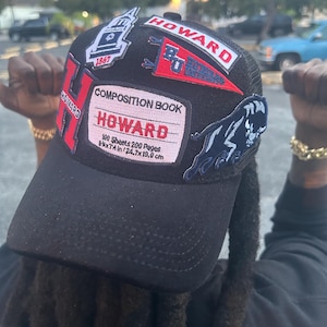May include: Black trucker hat with multiple patches. Patches include the word "HOWARD" in red and white, a composition book design, a panther, and the year "1867". The hat has a curved brim and a mesh back.
