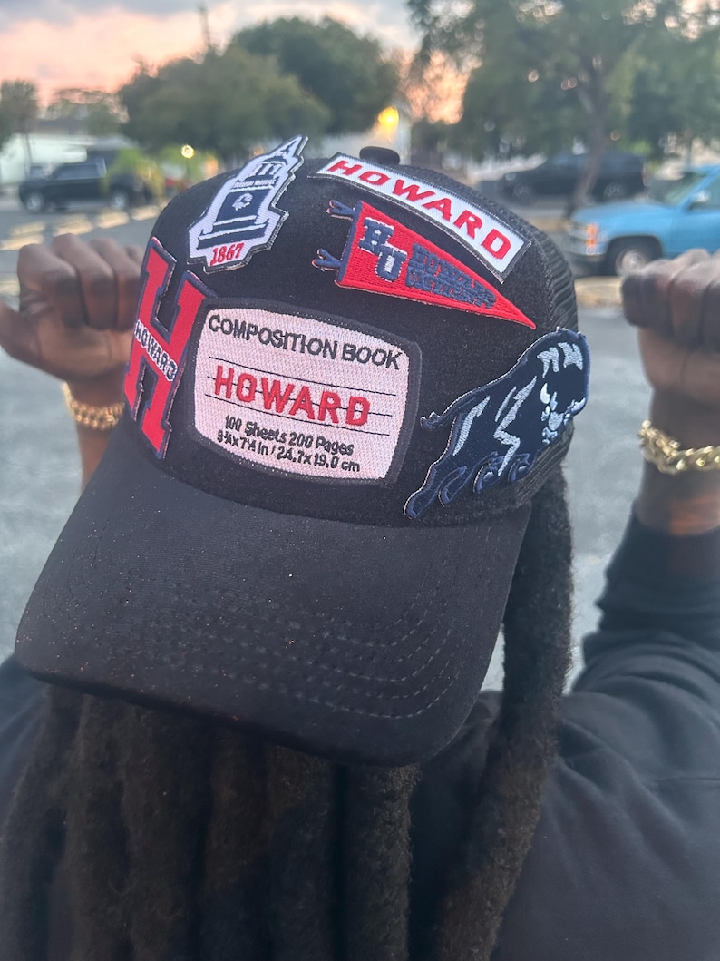 May include: Black trucker hat with multiple patches. Patches include the word "HOWARD" in red and white, a composition book design, a panther, and the year "1867". The hat has a curved brim and a mesh back.
