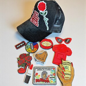 May include: A black trucker hat with a red rose patch and the text "I'M JUST A GIRL." Assorted embroidered patches include a red lipstick, a high-heeled shoe, and a credit card design. Other patches have text and graphic designs.