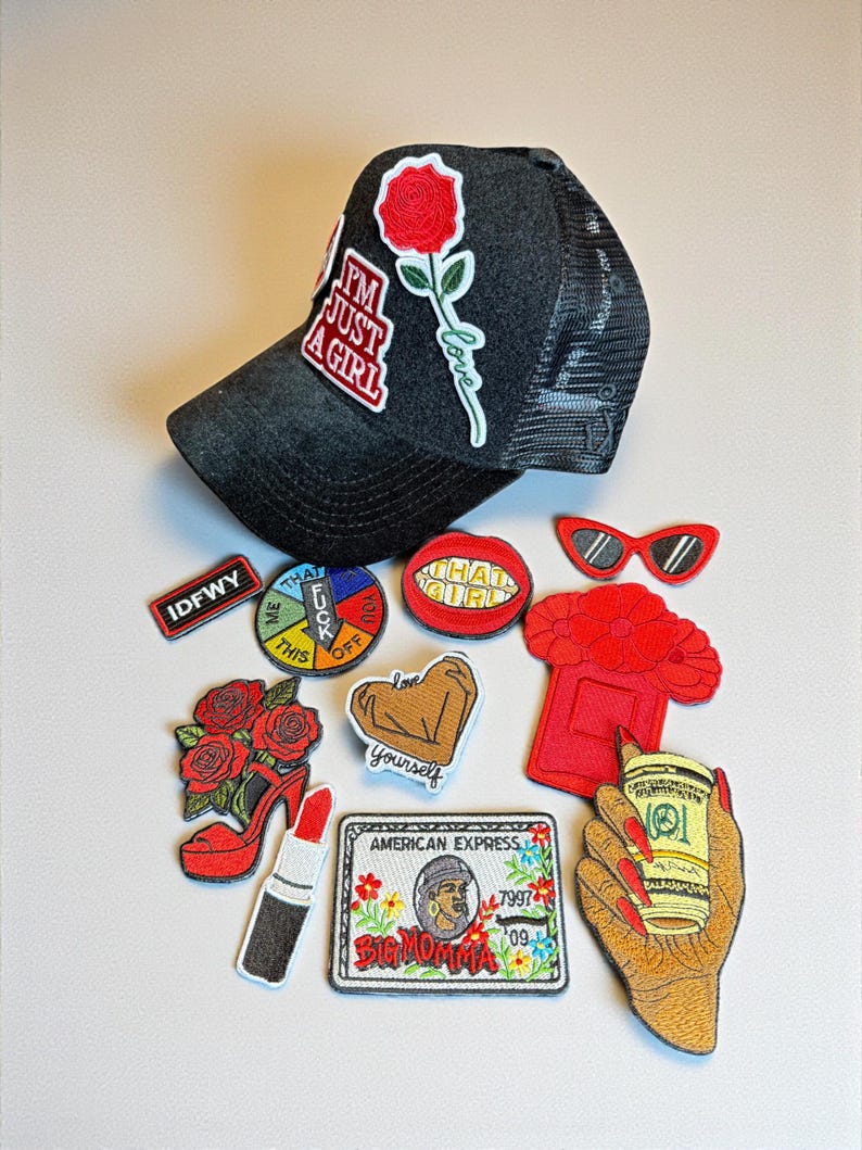 May include: A black trucker hat with a red rose patch and the text "I'M JUST A GIRL." Assorted embroidered patches include a red lipstick, a high-heeled shoe, and a credit card design. Other patches have text and graphic designs.