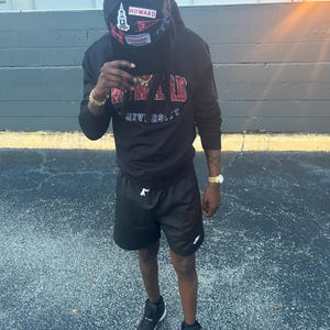 May include: A person wearing a black sweatshirt with "HOWARD UNIVERSITY" in red lettering, black shorts, and black sneakers with red accents. A black cap with "HOWARD" is worn.
