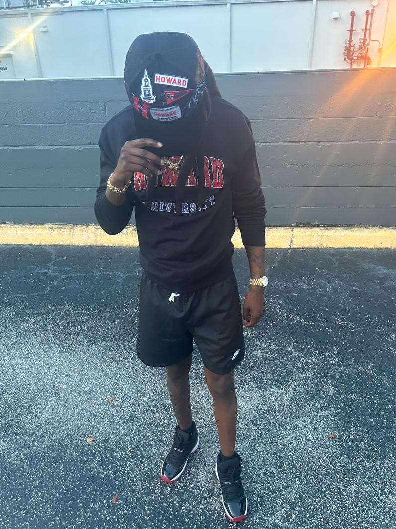May include: A person wearing a black sweatshirt with "HOWARD UNIVERSITY" in red lettering, black shorts, and black sneakers with red accents. A black cap with "HOWARD" is worn.