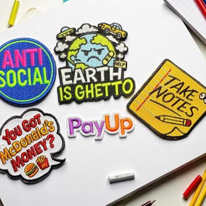Peut inclure: Plusieurs écussons brodés avec divers motifs et textes. L'un d'eux affiche "ANTI SOCIAL" en rose et vert sur fond bleu. D'autres écussons incluent "EARTH IS GHETTO", "TAKE NOTES", "You Got McDonald's MONEY?" et "PayUp".