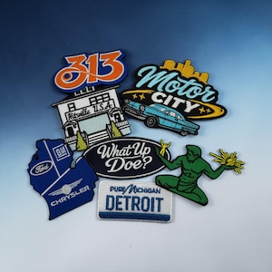 Op de afbeelding: Een verzameling geborduurde patches met verschillende ontwerpen en tekst. Patches zijn onder meer "313", "Motor City" met een auto, "Hitsville U.S.A.", een Michigan-omtrek met "Ford", "GM" en "Chrysler", "What Up Doe?", "Pure Michigan Detroit" en een groen figuur.