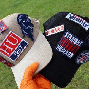 May include: Two baseball caps with patches. One is tan with a white brim, featuring a panther patch and "HU 1867" patch. The other is black with "HOWARD" and "STRAIGHT OUTTA HOWARD" patches. Both have multiple patches.