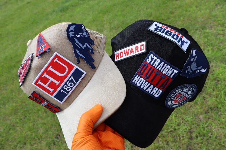 May include: Two baseball caps with patches. One is tan with a white brim, featuring a panther patch and "HU 1867" patch. The other is black with "HOWARD" and "STRAIGHT OUTTA HOWARD" patches. Both have multiple patches.