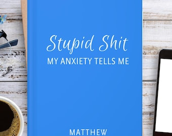 May include: A bright blue book with the white text "Stupid Shit MY ANXIETY TELLS ME" and "MATTHEW" printed on the front cover. The book is positioned next to a black smartphone with a blank white screen, a cup of black coffee, and black glasses.
