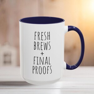 Peut inclure: Mug blanc avec intérieur et anse bleu marine. Le mug porte l'inscription noire "FRESH BREWS + FINAL PROOFS". Idéal pour les boissons chaudes, comme le café ou le thé.