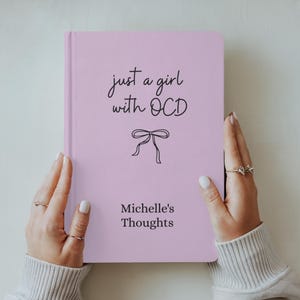May include: A pink journal with the text "just a girl with OCD" and "Michelle's Thoughts" in black script. A black bow graphic is centered below the first line of text. The journal is held by a person wearing rings.