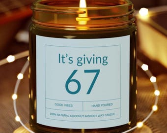 Puede incluir: Una vela encendida en un frasco de vidrio ámbar con una etiqueta azul claro que presenta el texto It's giving 67, GOOD VIBES, HAND POURED y 100% NATURAL COCONUT APRICOT WAX CANDLE. La vela tiene una mecha encendida con una llama visible arriba.