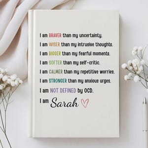 May include: A white journal with the text "I am BRAVER than my uncertainty" and other affirmations in various colors. The journal is personalized with the name "Sarah" and a heart symbol.