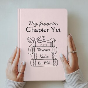 May include: A pale pink journal featuring the text "My favourite Chapter Yet" and a stack of books with "30 years", "Katie", and "Est. 1996" printed on them. The journal is held by two hands with rings.