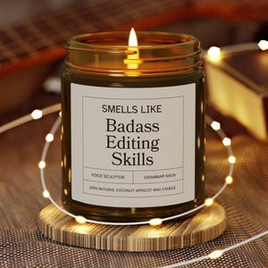 Puede incluir: Una vela encendida en un tarro de cristal con una etiqueta que dice "Smells Like Badass Editing Skills". La vela está sobre un posavasos de madera y está rodeada de luces de cadena. La etiqueta también dice "Voice Sculptor" y "Grammar Ninja".
