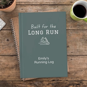 May include: A teal journal with the text "Built for the LONG RUN" and a white line drawing of a running shoe. Below the text is "Emily's Running Log". The journal is on a wooden surface with a spiral notebook and a cup of coffee.