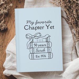 May include: A light blue book with the text "My favorite Chapter Yet" and a stack of books with a bow illustration. The books read "50 years", "Sarah", and "Est. 1976". The book is on a light-colored cloth.