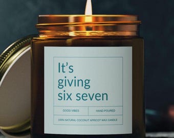Puede incluir: Una vela encendida en un frasco de vidrio ámbar con tapa dorada. La etiqueta blanca presenta el texto en color verde azulado It's giving six seven, Good Vibes, Hand Poured y 100% Natural Coconut Apricot Wax Candle en un borde rectangular.