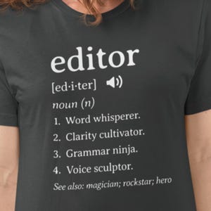 May include: A dark grey t-shirt with white text that reads "editor" and a definition of the word. The definition includes the following: "Word whisperer, Clarity cultivator, Grammar ninja, Voice sculptor. See also: magician; rockstar; hero"