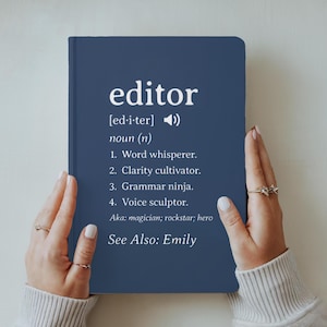 May include: A navy blue notebook with the word "editor" in white, along with a definition. The definition includes terms like "Word whisperer" and "Grammar ninja." The notebook is held by hands with rings.