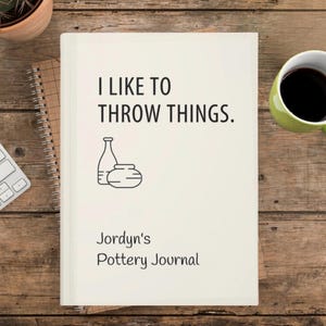 May include: A cream journal with the text "I LIKE TO THROW THINGS." and "Jordyn's Pottery Journal" in black. The cover features a line drawing of a bottle and a bowl. The journal is on a wooden surface.