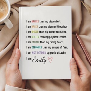 May include: A white journal with black and colored text, held by hands. The text reads, "I am BRAVER...WISER...BIGGER...SOFTER...CALMER...STRONGER...NOT DEFINED...I am Emily." A pink pen and a cup of coffee are in the background.