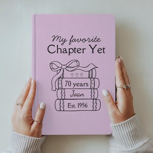 May include: A pale pink journal featuring the text "My favourite Chapter Yet" and a stack of books with "70 years", "Joan", and "Est. 1956" printed on them. The journal is held by a person.