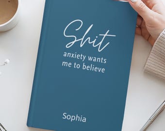 May include: A teal journal with the word "Shit" in cursive script, followed by "anxiety wants me to believe." The name "Sophia" is printed at the bottom. A hand holds the journal near a cup of coffee and a pen.