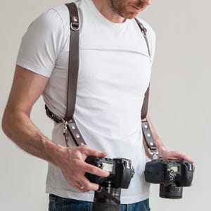 Puede incluir: Arnés doble de cuero marrón para cámara con herrajes plateados. El arnés está diseñado para sujetar dos cámaras, con correas ajustables y clips metálicos. El fotógrafo lleva una camiseta blanca y vaqueros azules.