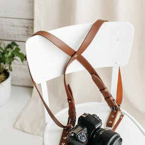 Puede incluir: Una cámara negra con lente descansa sobre una silla blanca. Una correa de cuero marrón está unida a la cámara. La correa está diseñada para usarse sobre ambos hombros. La cámara es una Nikon.