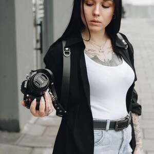 May include: A black Nikon camera with a black lens cap is held by a person. The camera has a black leather strap. The person is wearing a black jacket over a white tank top and blue jeans. The person has tattoos on their arms.