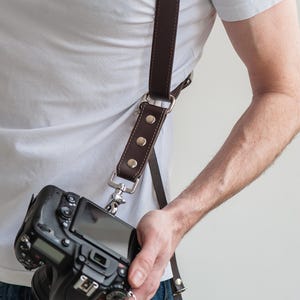 May include: A brown leather camera strap with silver metal hardware and rivets. The strap is worn over the shoulder and attaches to a black DSLR camera. The camera is held in the hand.