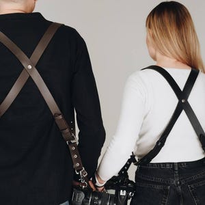 Puede incluir: Dos personas con correas de cuero para cámara, sosteniendo cámaras. Las correas son marrones y negras, se cruzan sobre los hombros y se sujetan a las cámaras. La persona de la izquierda lleva una camiseta negra de manga larga y vaqueros. La persona de la derecha lleva una camiseta blanca de manga larga y vaqueros negros.