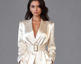 May include: A champagne-colored satin suit consisting of a long-sleeved blazer with a notched lapel and a matching belt with a gold buckle, paired with wide-leg trousers. The fabric has a smooth, reflective finish and a tailored fit.