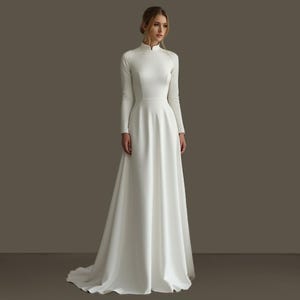 May include: A long, white wedding dress with a high neckline and long sleeves. The dress has a fitted bodice and a flowing A-line skirt. The fabric appears to be a smooth, matte material. The dress is floor-length and has a slight train.