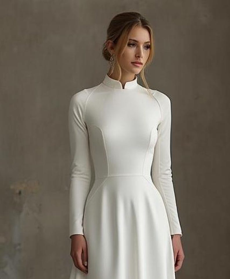 May include: A long-sleeved, white wedding dress with a high neckline and a flared skirt. The dress features a fitted bodice and a simple, elegant design. The dress is made of a smooth, opaque fabric.