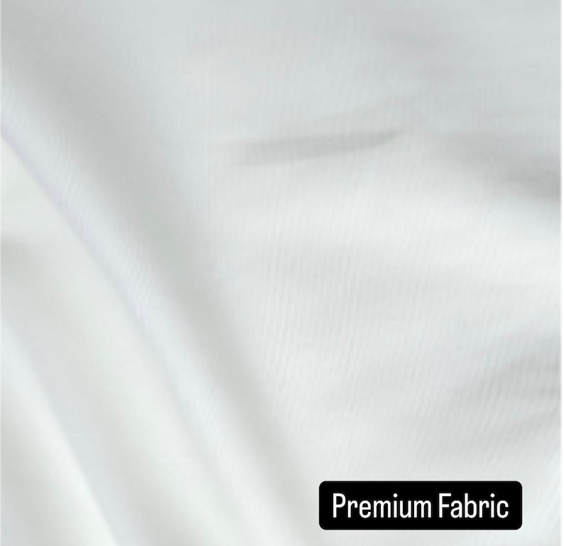 May include: Close-up of white fabric with subtle diagonal texture. The fabric is draped, creating soft folds and shadows. The text "Premium Fabric" is displayed in a black rectangle at the bottom of the image. The fabric appears to be smooth and high-quality.