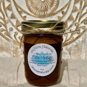 May include: A glass jar with a gold lid and a brown label that reads "Ja'Nova Naturals Fire Honey 1-2 tsp in warm water Organic Ingredients: Honey, Cinnamon, Cardamom, Turmeric, Ginger, Cayenne, Black Pepper 8oz. janovanaturals.com".
